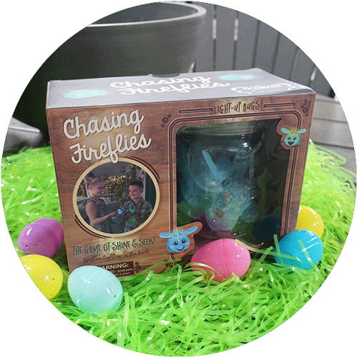 The packaged Chasing Fireflies Shine & Seek Game, laying on green easter grass with colorful eggs around it.